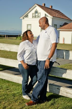 A couple in matching white shirts and jeans smiling at each other by a white fence with a house behind them.