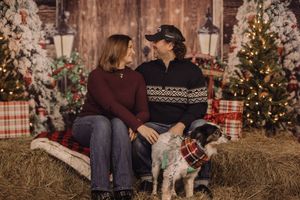 Couple sitting on hay bale with dog in front of Christmas trees and festive holiday decorations.