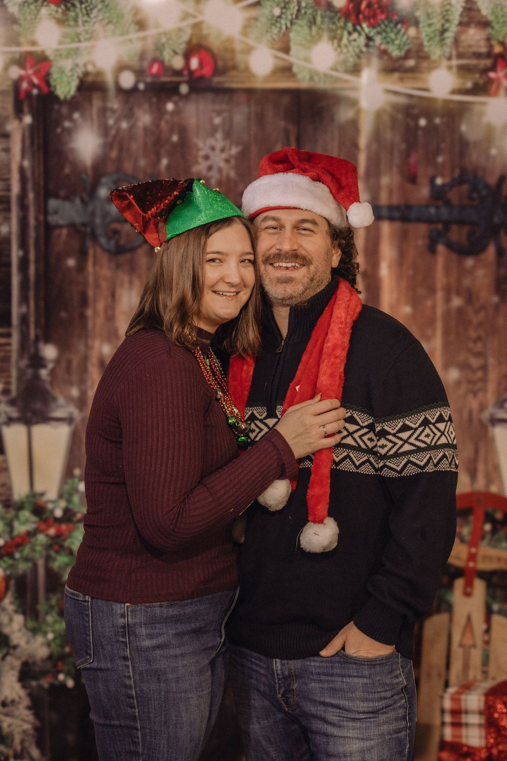 Couple smiling in Christmas hats posed before festive wooden backdrop with holiday lights and greenery.