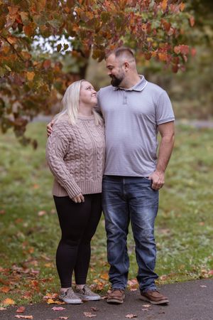 A couple in casual attire stands together on a pathway surrounded by autumn trees with red leaves.