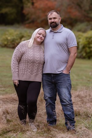 A couple stands together in a field during autumn wearing a beige cable knit sweater and casual clothing.