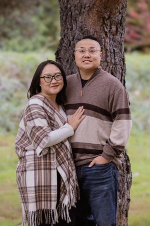 A couple in coordinating brown outfits poses together against a tree trunk in an outdoor setting.