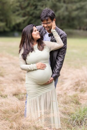 A couple embraces lovingly in a field of tall grass while wearing a cream dress and black jacket.