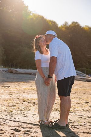 Couple sharing a romantic kiss on a sunny beach, woman smiling in striped skirt, man in white shirt and cap.