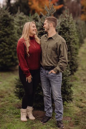 Couple smiling at each other at a Christmas tree farm, woman in red sweater, man in olive shirt and jeans.