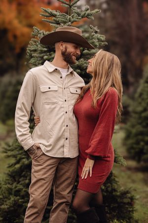 A couple in casual fall attire shares intimate moments among evergreen trees at a Christmas tree farm.