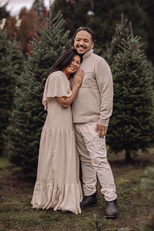 A couple shares an intimate moment among evergreen trees at a Christmas tree farm wearing coordinated neutral outfits.