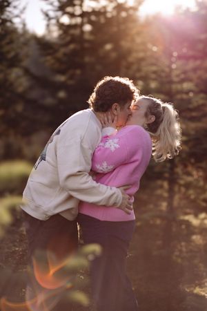 A romantic couple embraces and kisses in a sunlit outdoor setting with golden light filtering through trees.