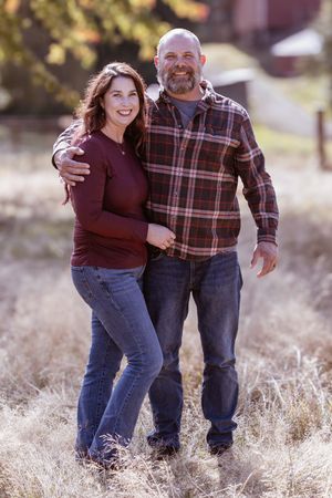 A couple wearing plaid and burgundy laughs together while walking through a golden field at sunset.