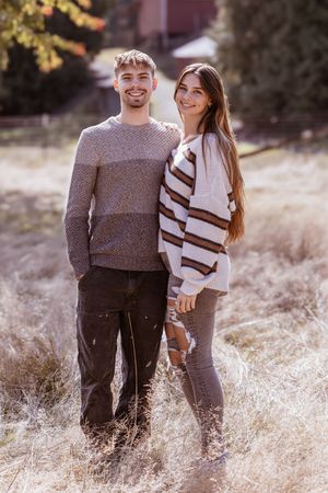 A couple wearing cozy sweaters embraces while standing in a sun-lit field of tall dried grass.