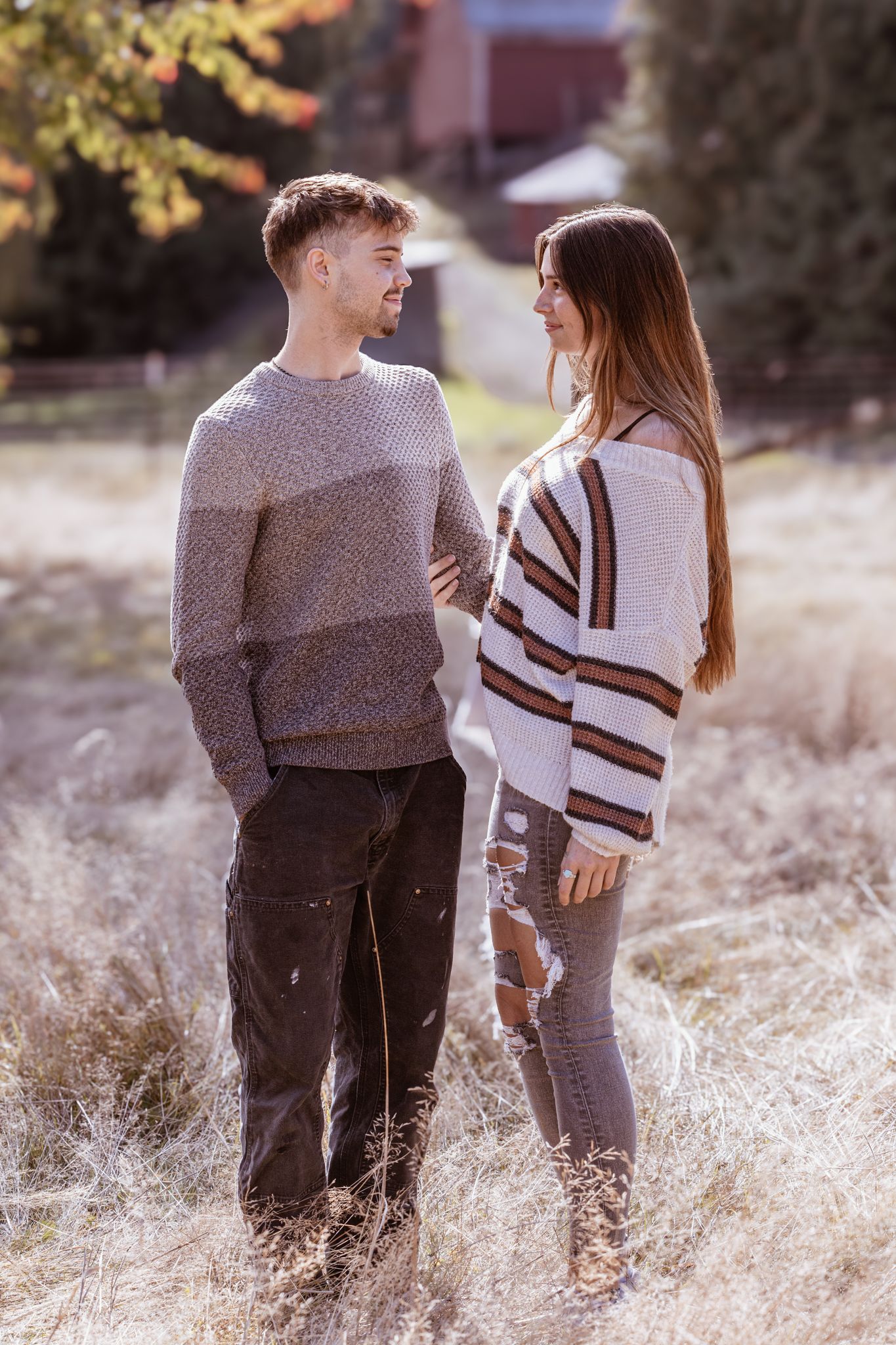 A couple wearing matching sweaters stands together in a field of tall dried grass at sunset.