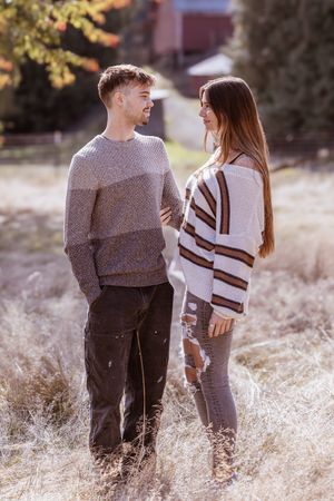 A couple wearing matching sweaters stands together in a field of tall dried grass at sunset.