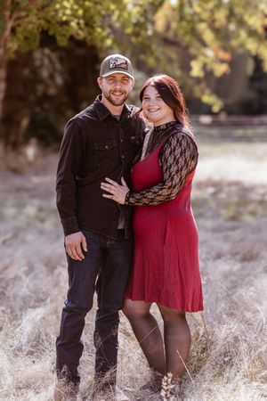 A couple poses together in a field during autumn, with the woman wearing a red dress and black lace sleeves.