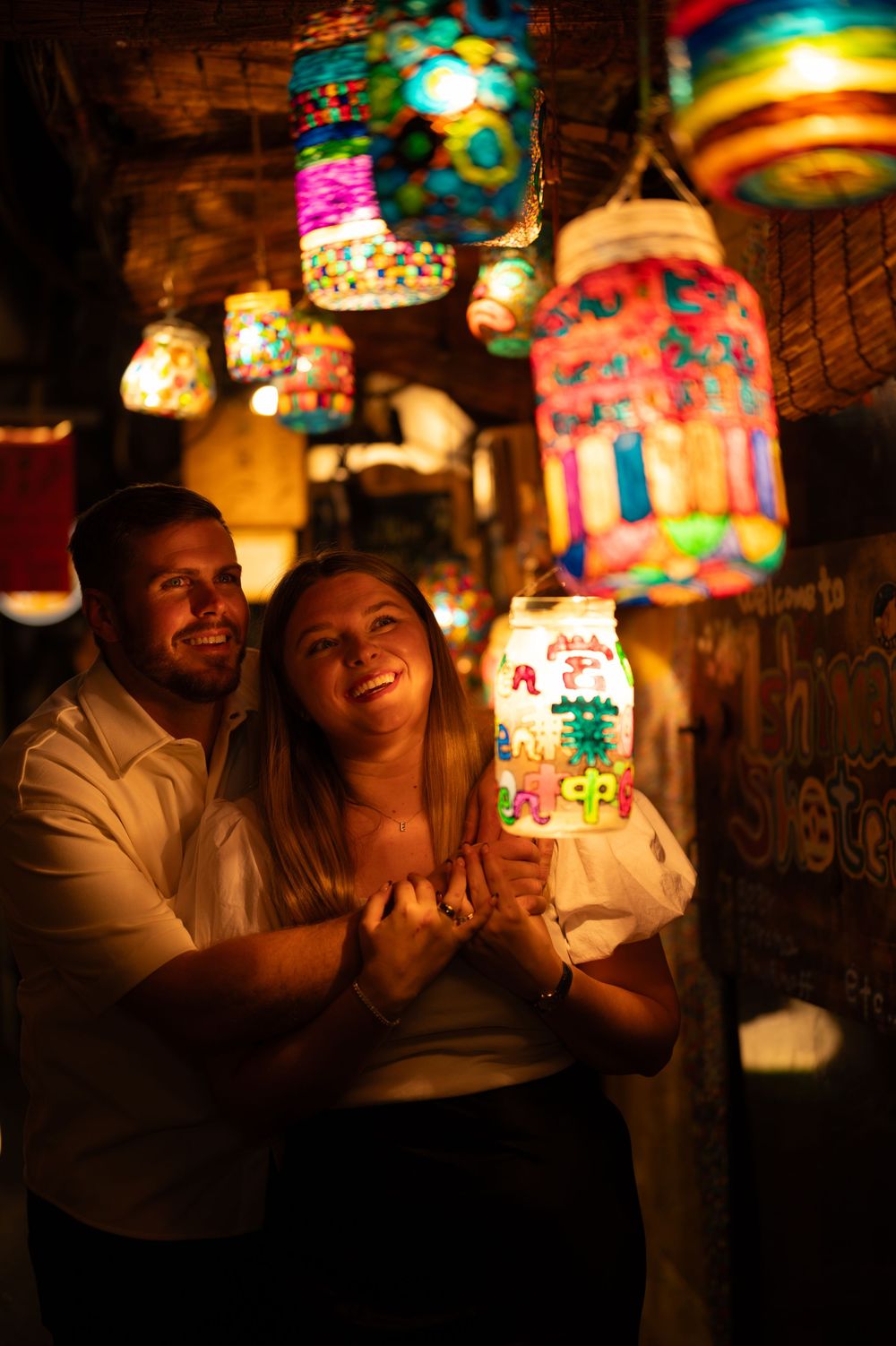 Happy couple holding a colorful illuminated lantern surrounded by vibrant hanging lanterns at night.