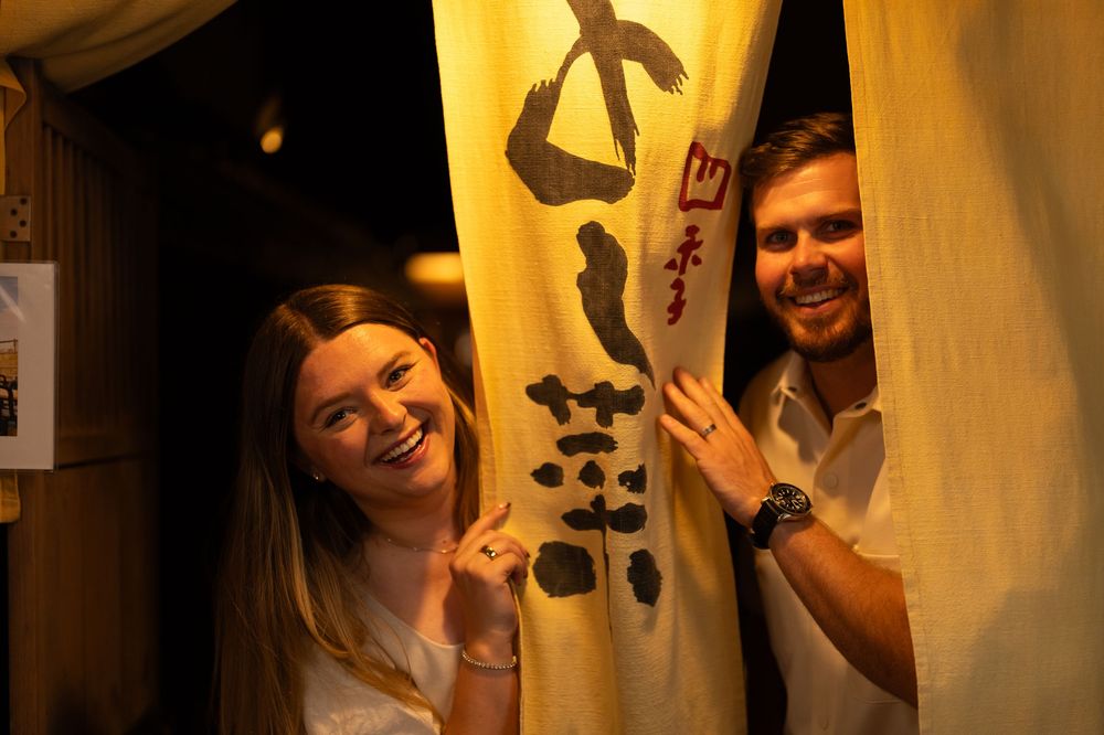 Smiling couple peeking playfully from behind a traditional Japanese noren curtain at a restaurant.