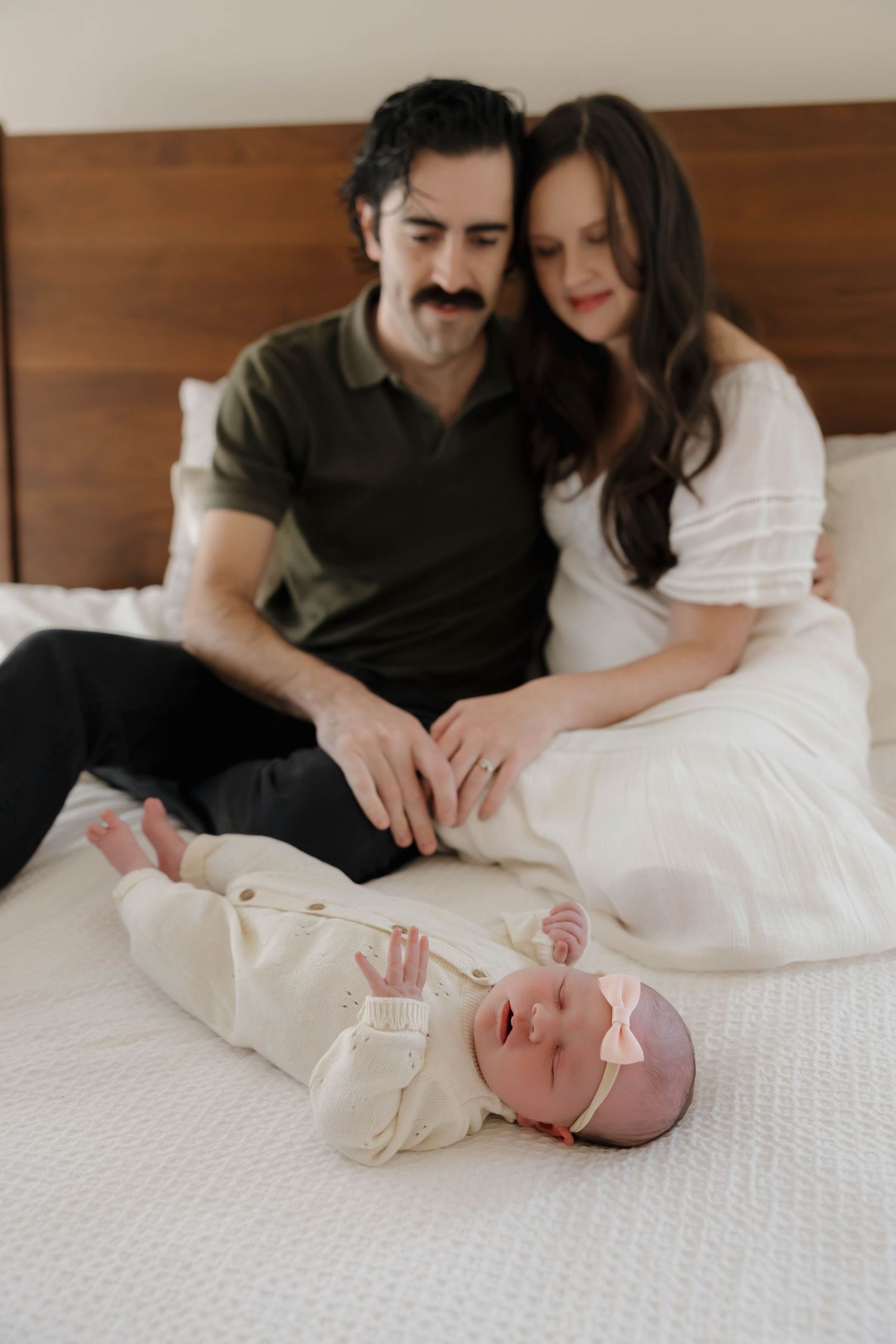 Parents lovingly watch their newborn baby lying on white bedding during an intimate lifestyle photography session.