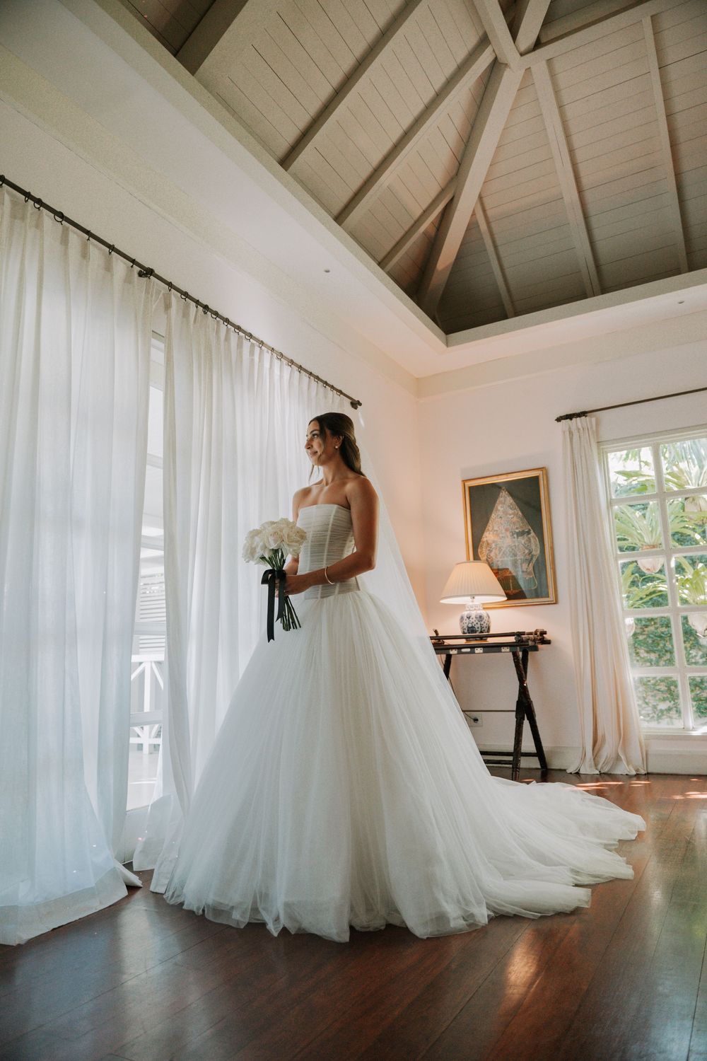 A bride in a flowing white ballgown twirls elegantly in a bright sunlit room with white curtains and hardwood floors.