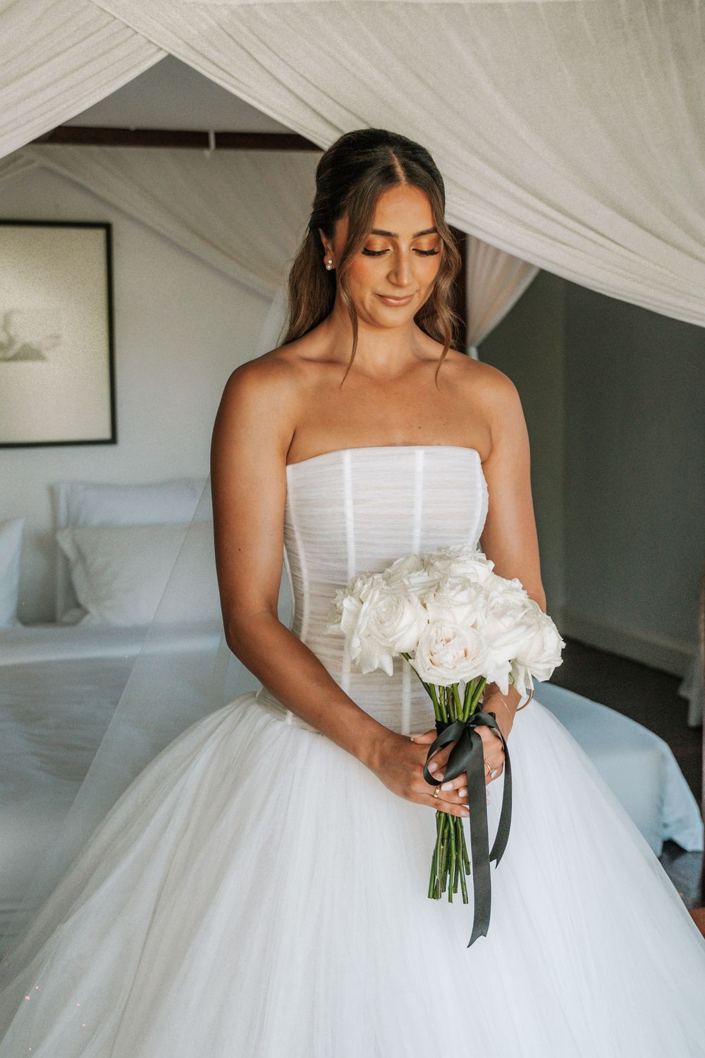 Bride in strapless white ball gown holding elegant bouquet of white peonies with black ribbon in sunlit bedroom.