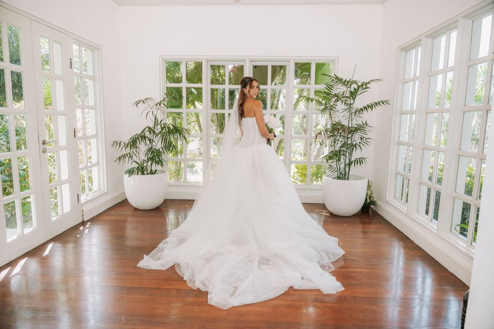 A panoramic view of a sunlit bridal suite with hardwood floors, white windows and a flowing wedding dress.