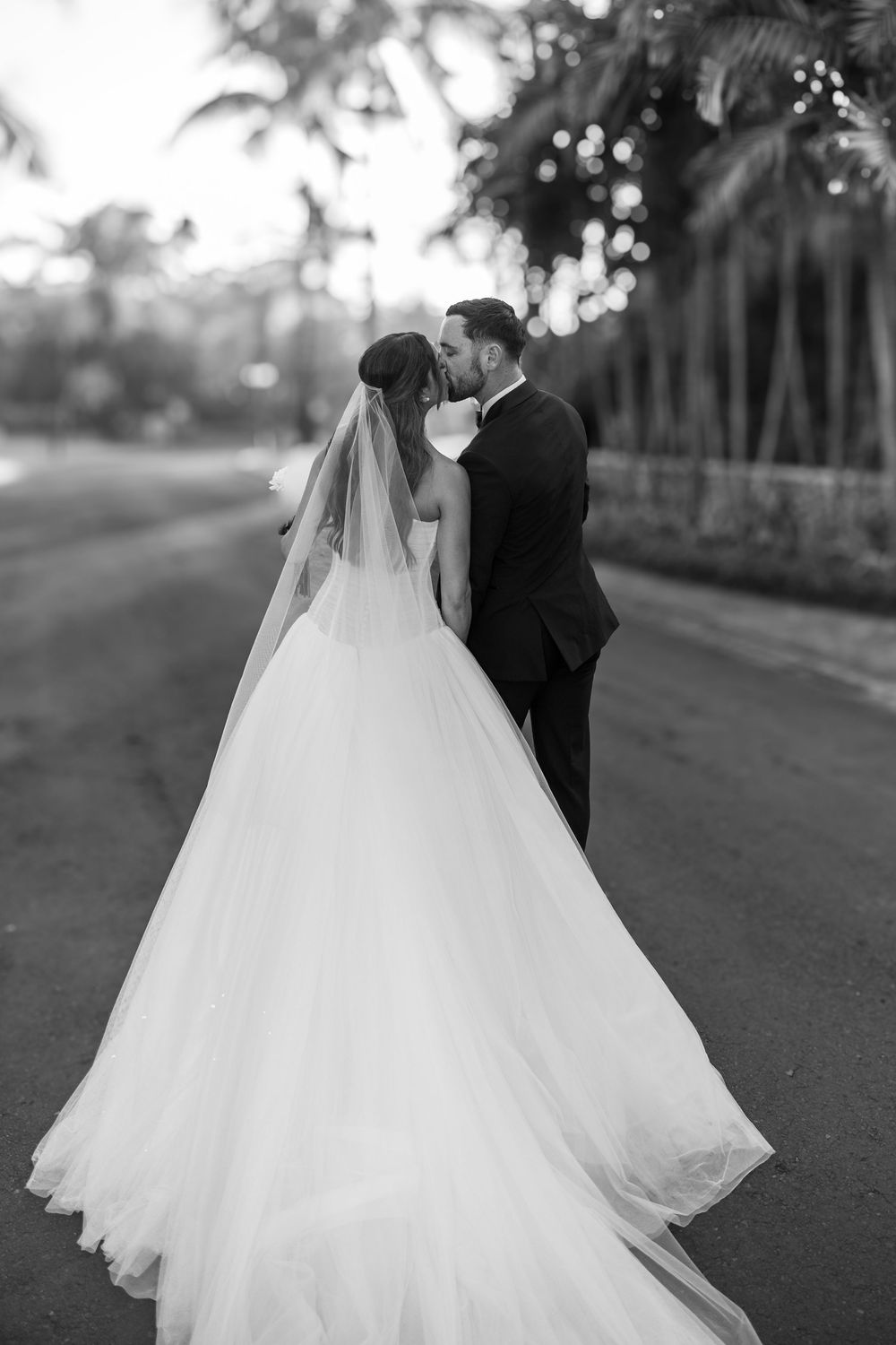 A couple in wedding attire embraces on a tree-lined path in a black and white romantic photograph.