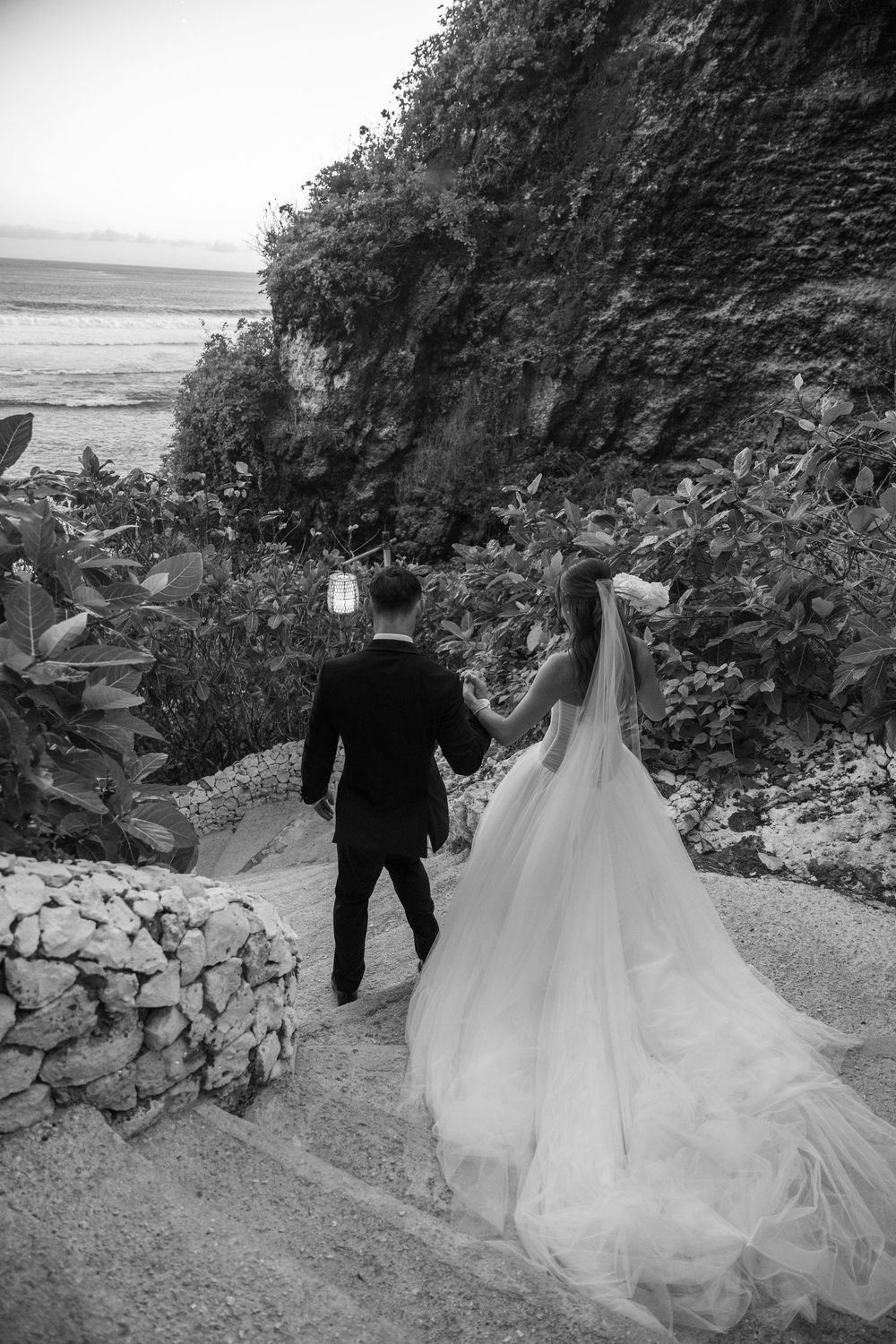 A couple in wedding attire walk down a scenic stone-lined path surrounded by lush greenery and cliffs.