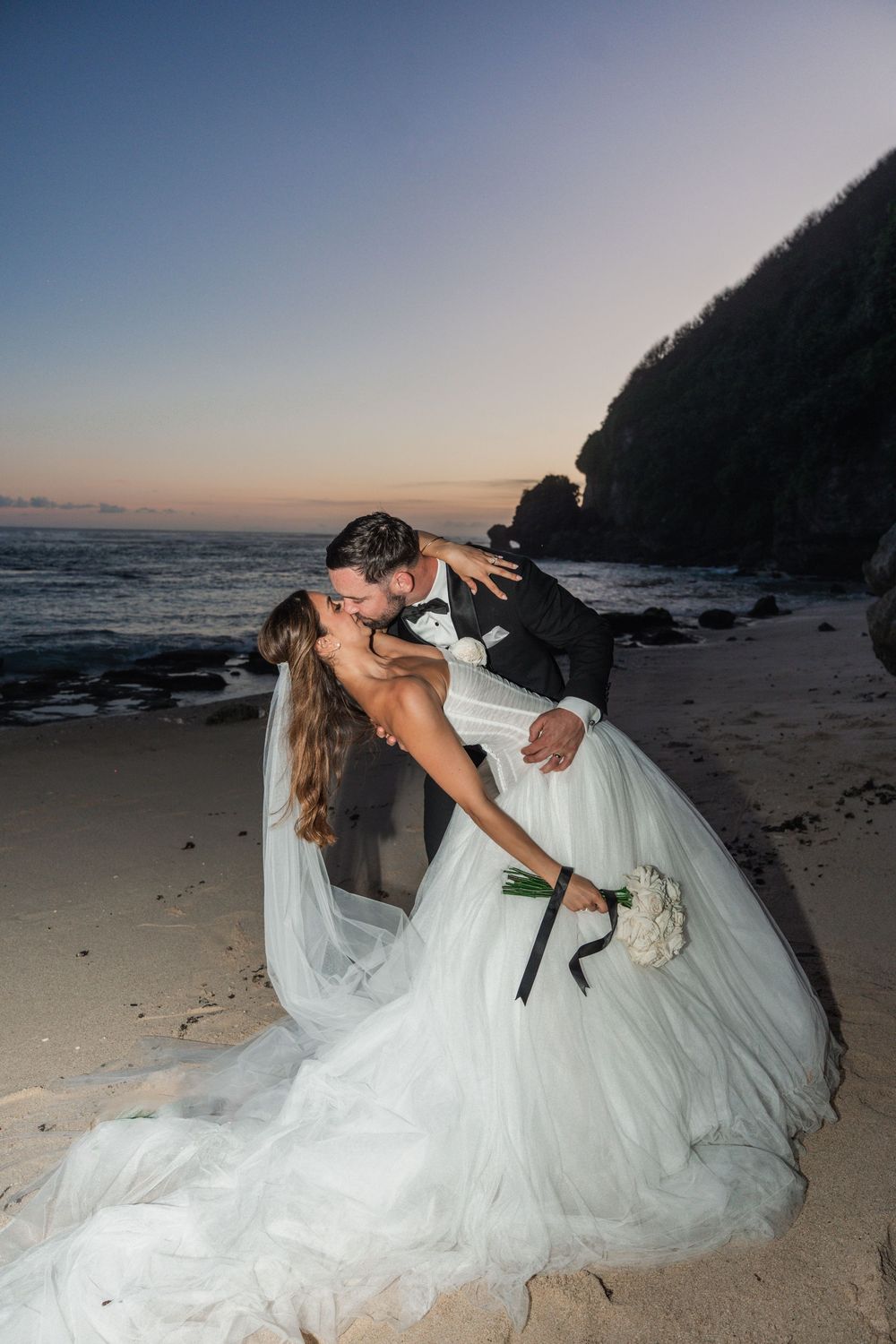 A romantic beach wedding photo series showing a couple in a dramatic dip pose at sunset by the ocean.