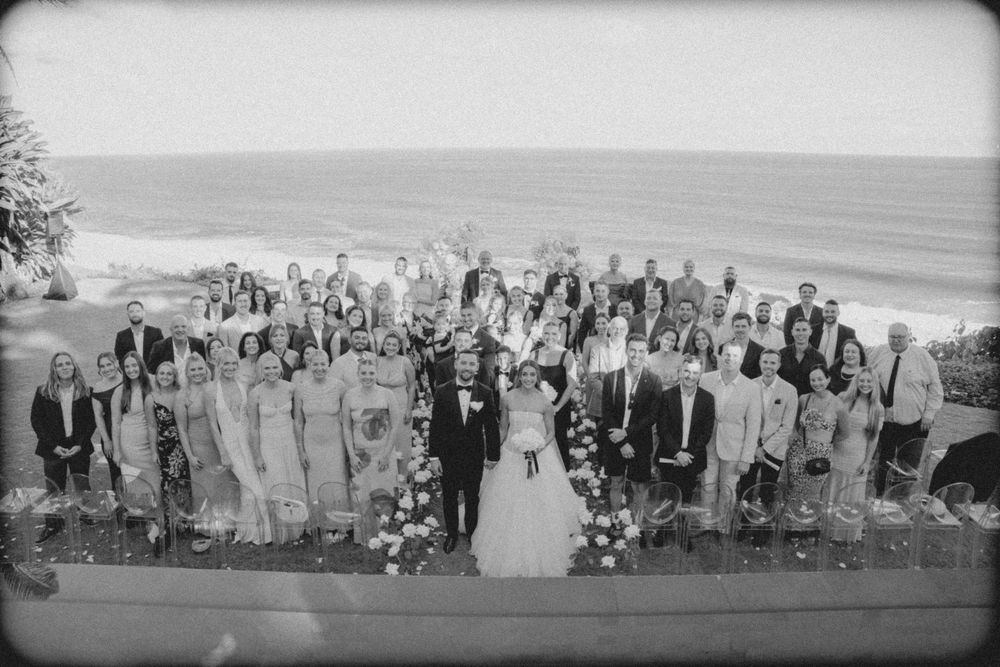 Black and white panoramic photo showing a large wedding party gathered on a beach with ocean waves in the background.