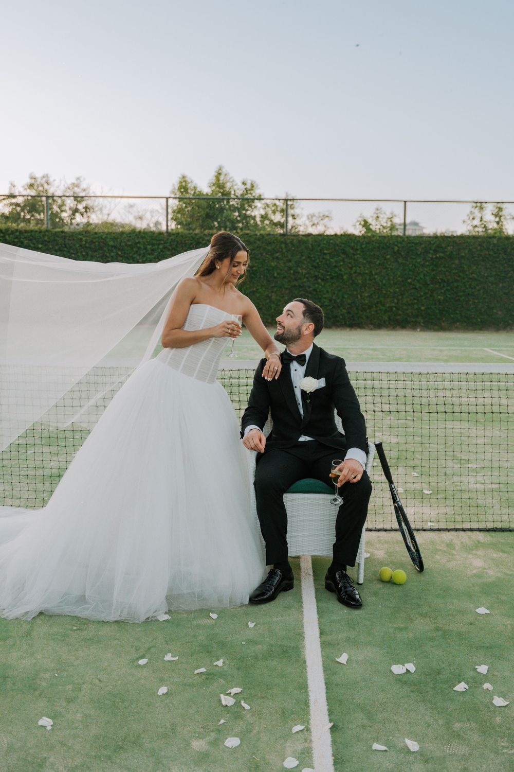 Newlyweds share a playful moment on a tennis court with scattered flower petals on the grass.