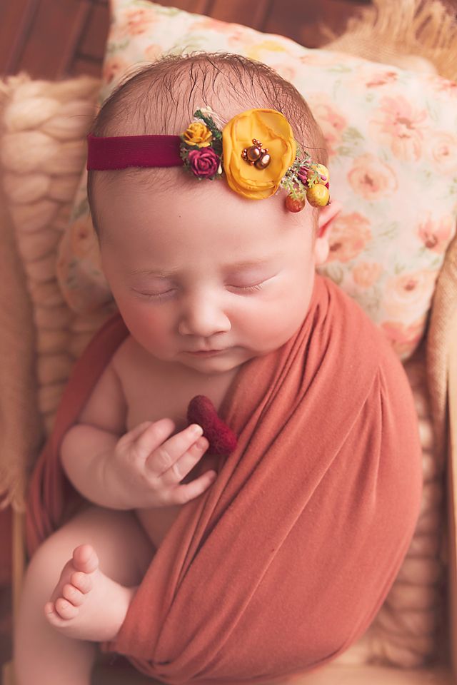 A sleeping newborn wrapped in a rust-colored swaddle wearing a delicate floral headband in warm autumn tones.