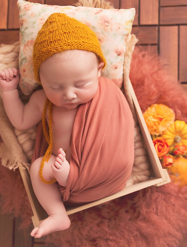 A sleeping baby in a peach outfit and yellow knit hat lies on soft pink tulle fabric next to wooden frames.