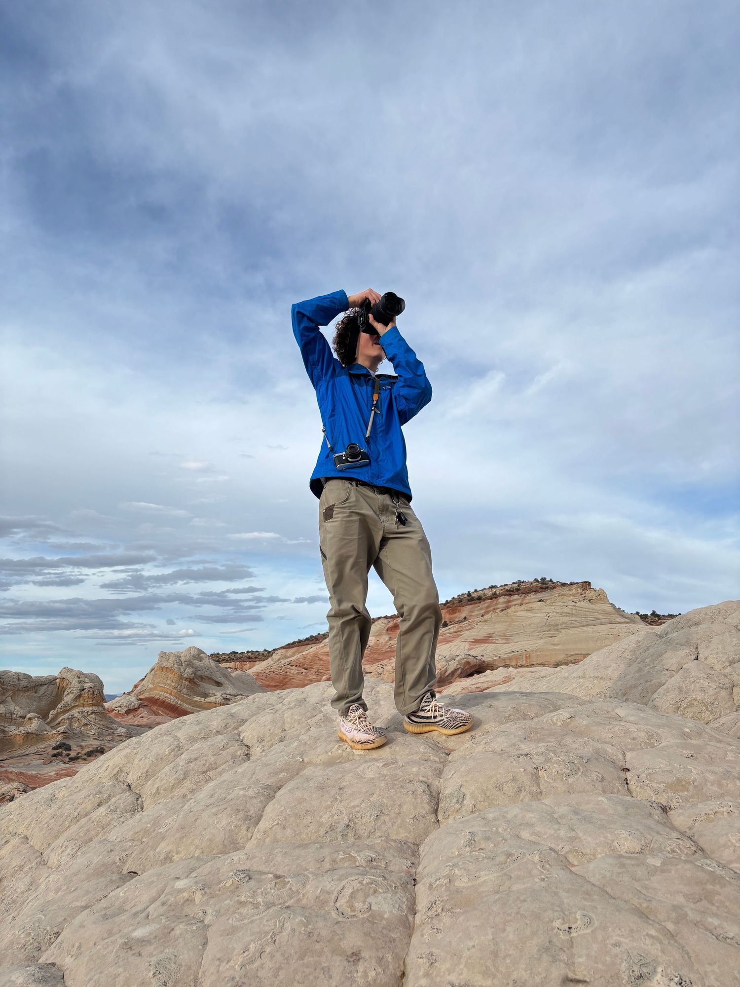 Hiker in blue jacket and khaki pants stands on red rock formations looking through binoculars against cloudy sky.