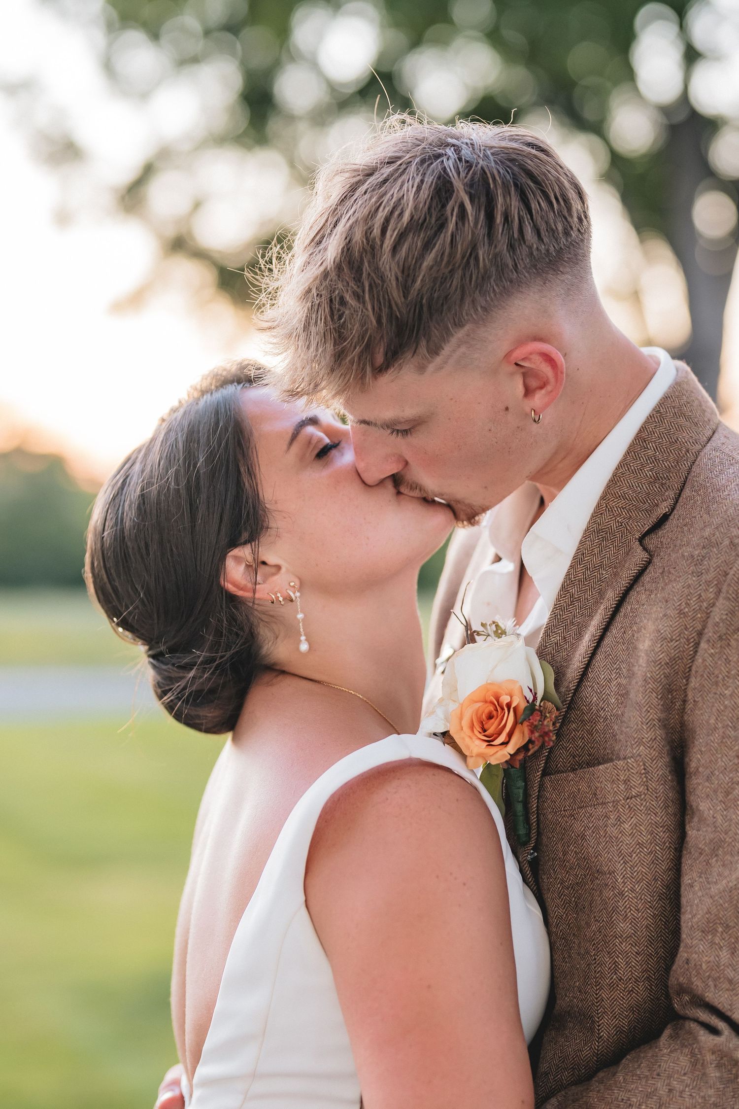 A romantic wedding couple shares tender kisses in a series of outdoor photos with a brown suit and peach boutonniere.