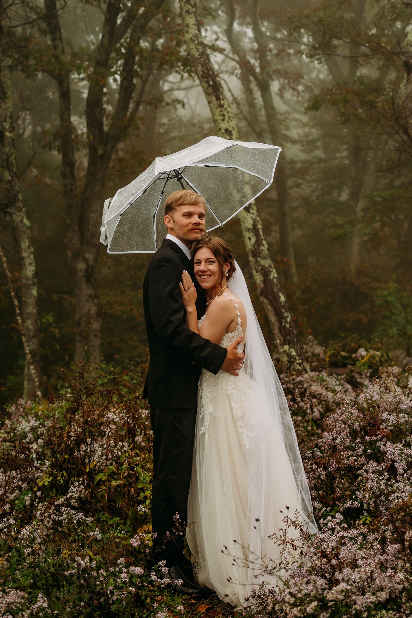 Picture of couple among flowers and trees in the background. Male is holding umbrella.