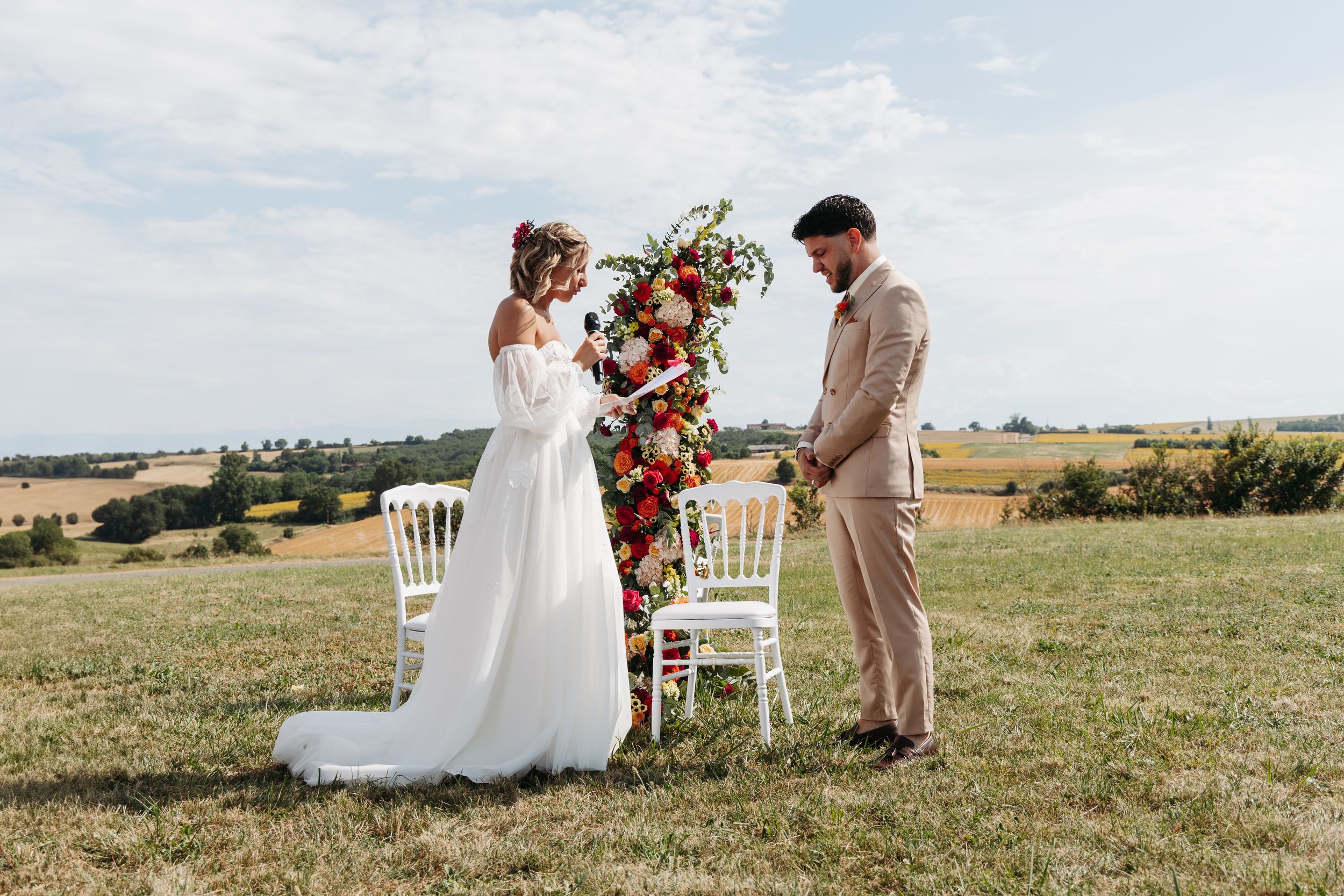 Bride and groom exchange vows outdoors at a floral arch ceremony in a sunlit countryside field.