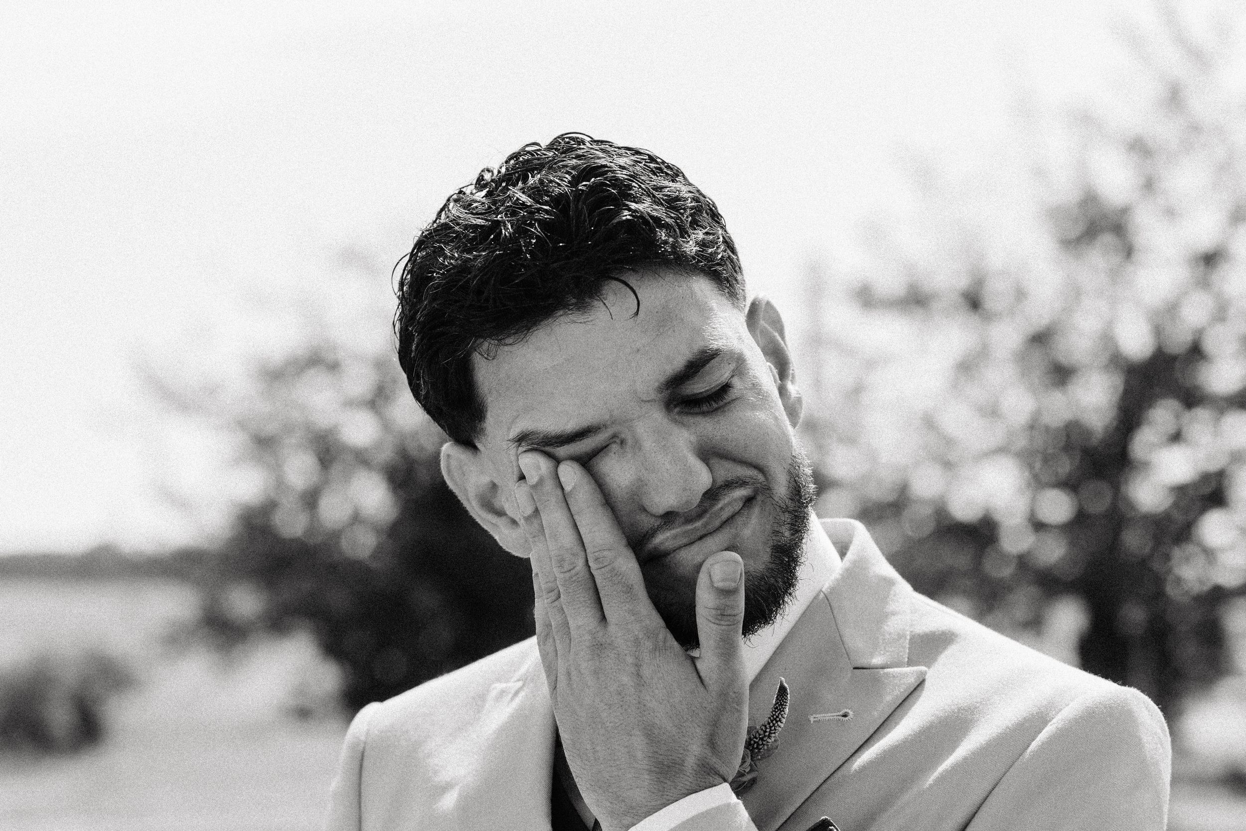 Emotional groom wiping tears during outdoor wedding ceremony in black and white portrait photo.