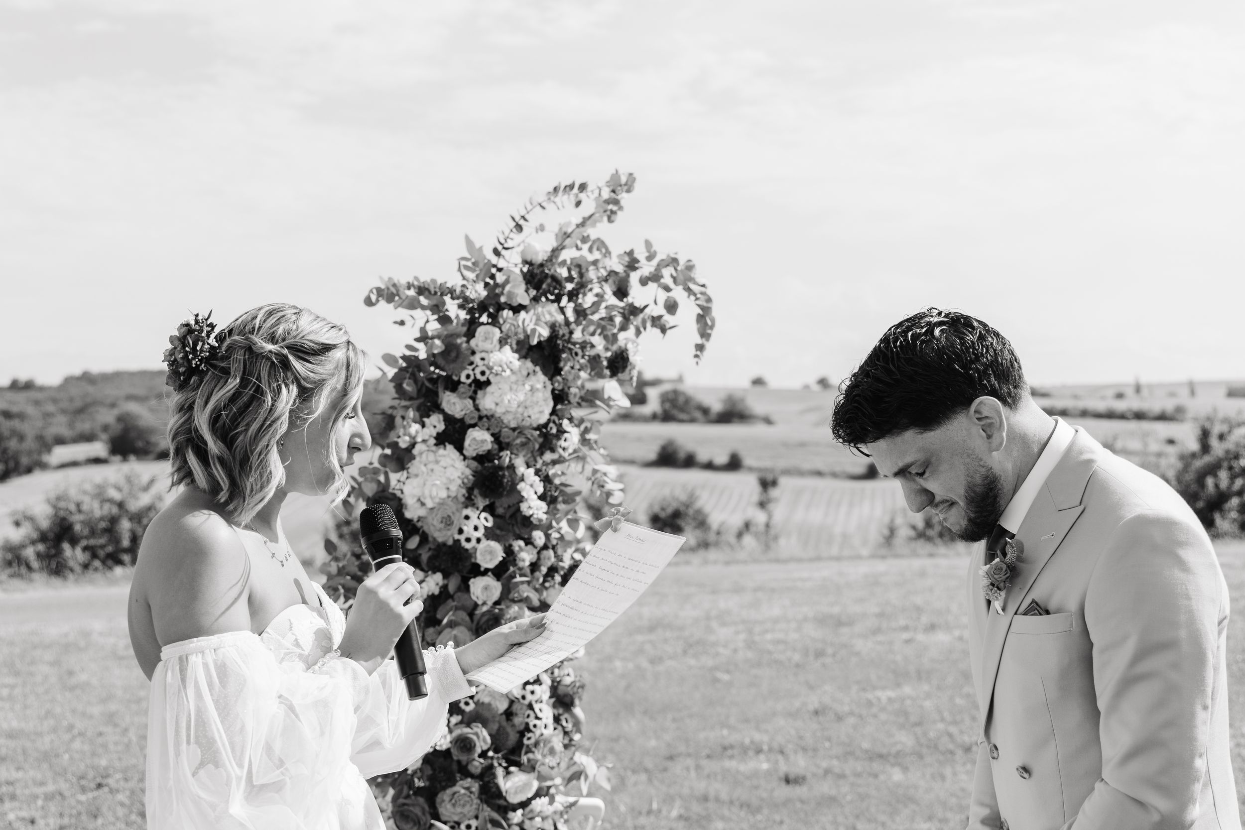 Bride reads wedding vows to groom at floral arch in black and white outdoor ceremony photo.