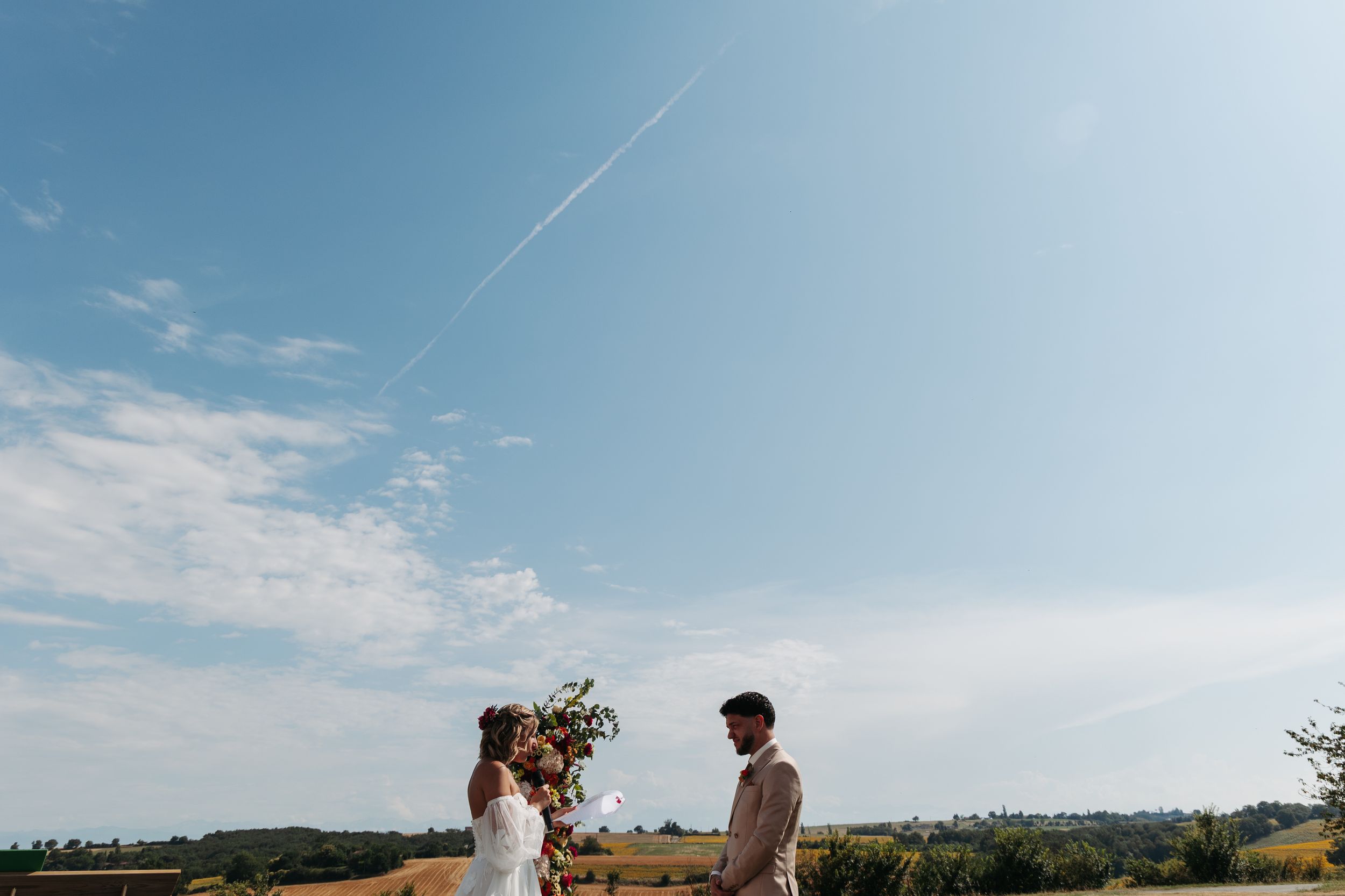 Bride and groom stand facing each other under a vast blue sky in an open countryside setting.