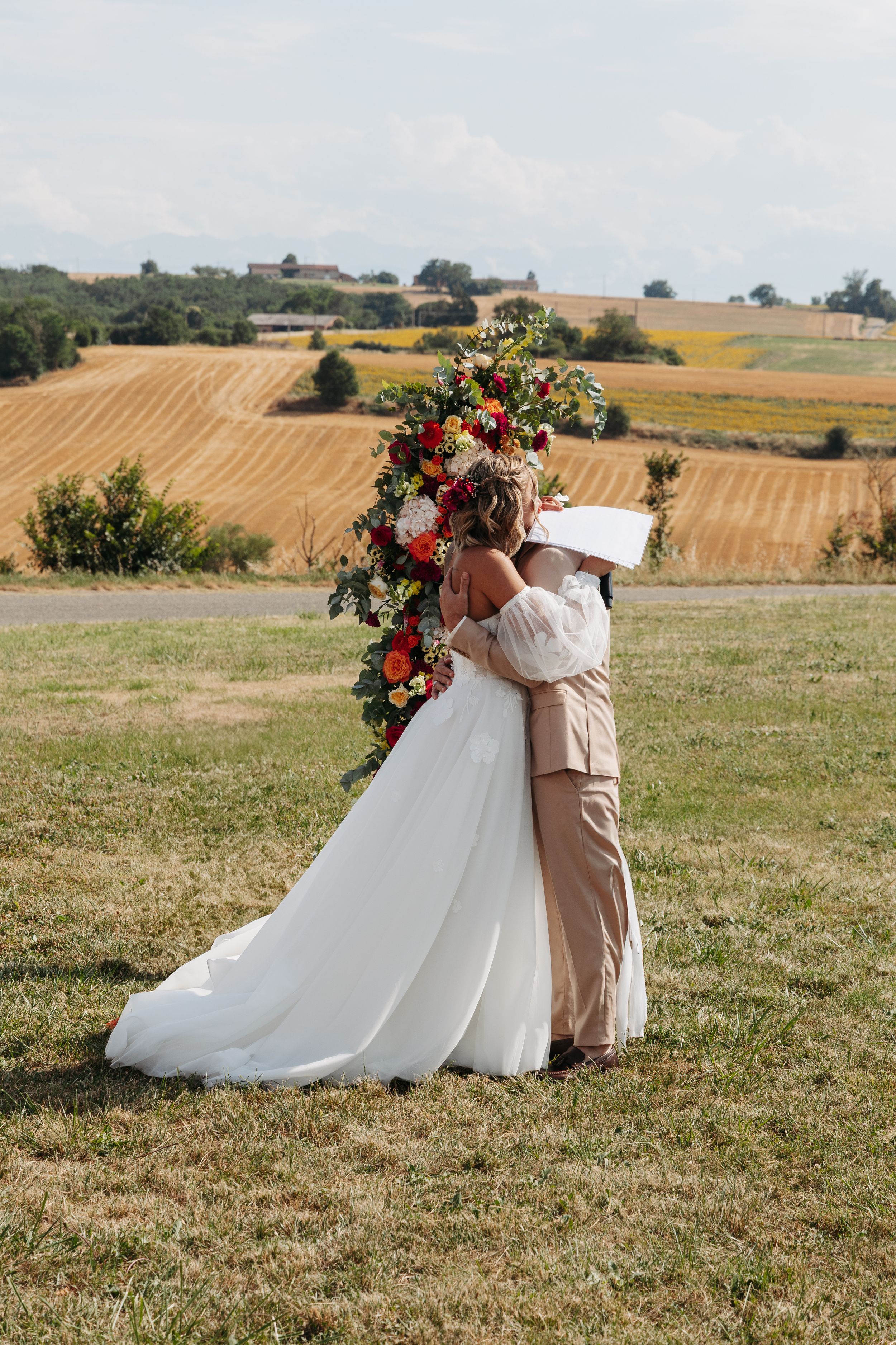 Newlyweds share a tender embrace beside a vibrant floral arch with rolling farmland backdrop.