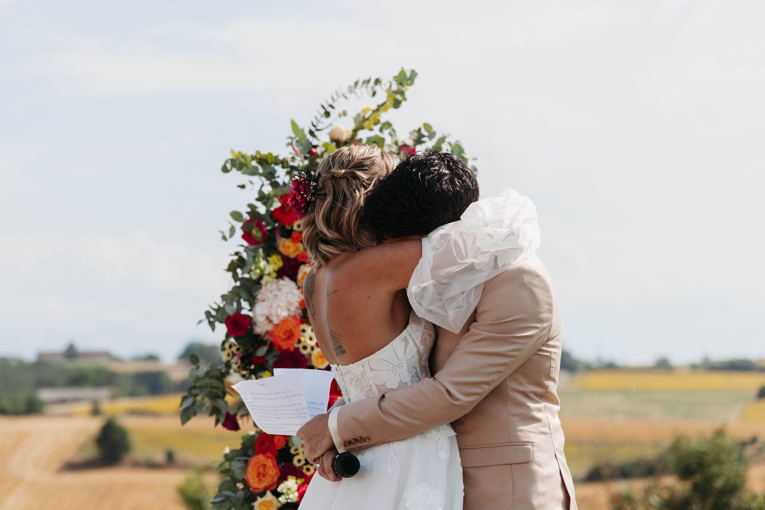Bride and groom share first kiss surrounded by colorful floral arch at outdoor countryside wedding.