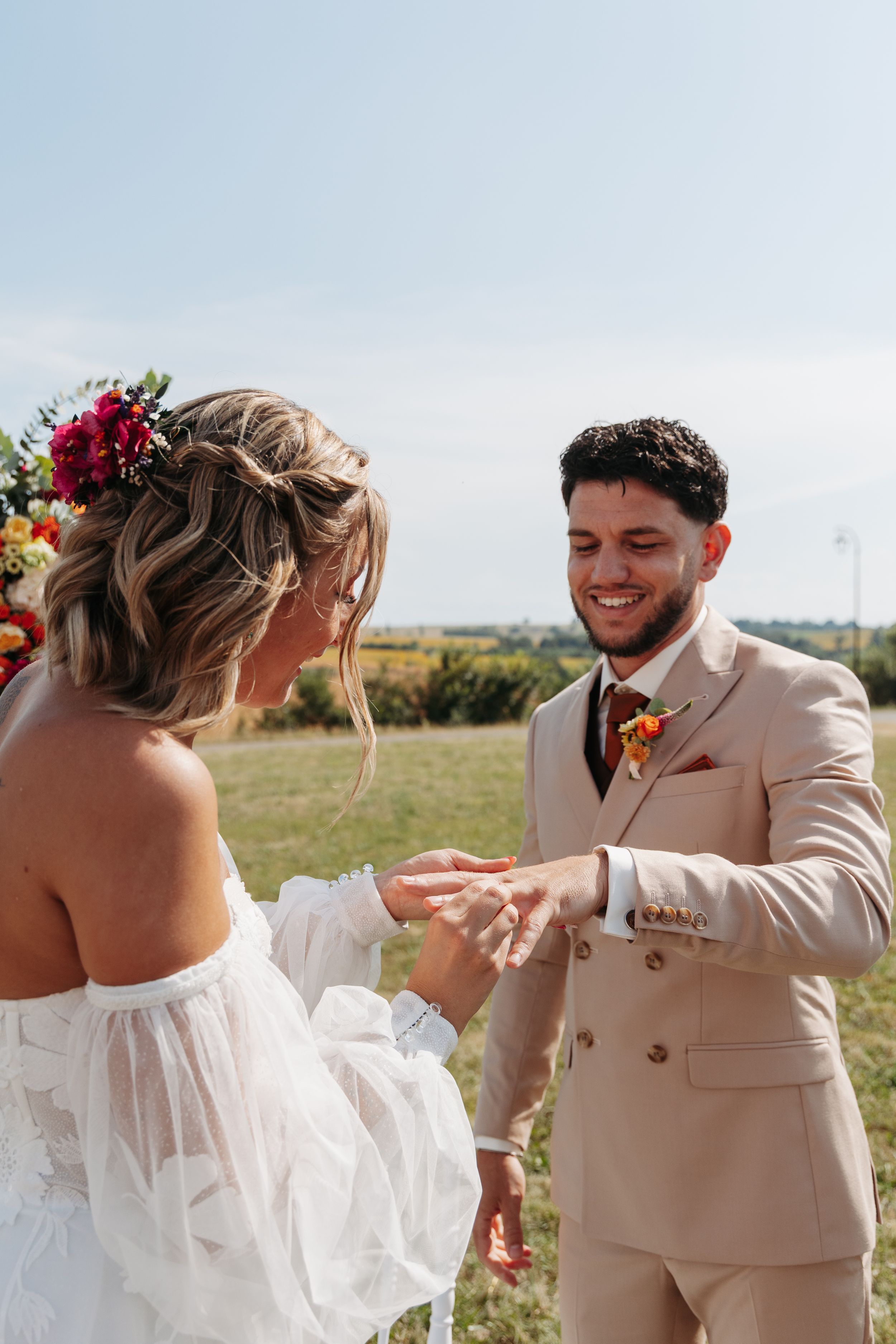 Smiling groom receives wedding ring from bride during sunny outdoor ceremony in beige suit.