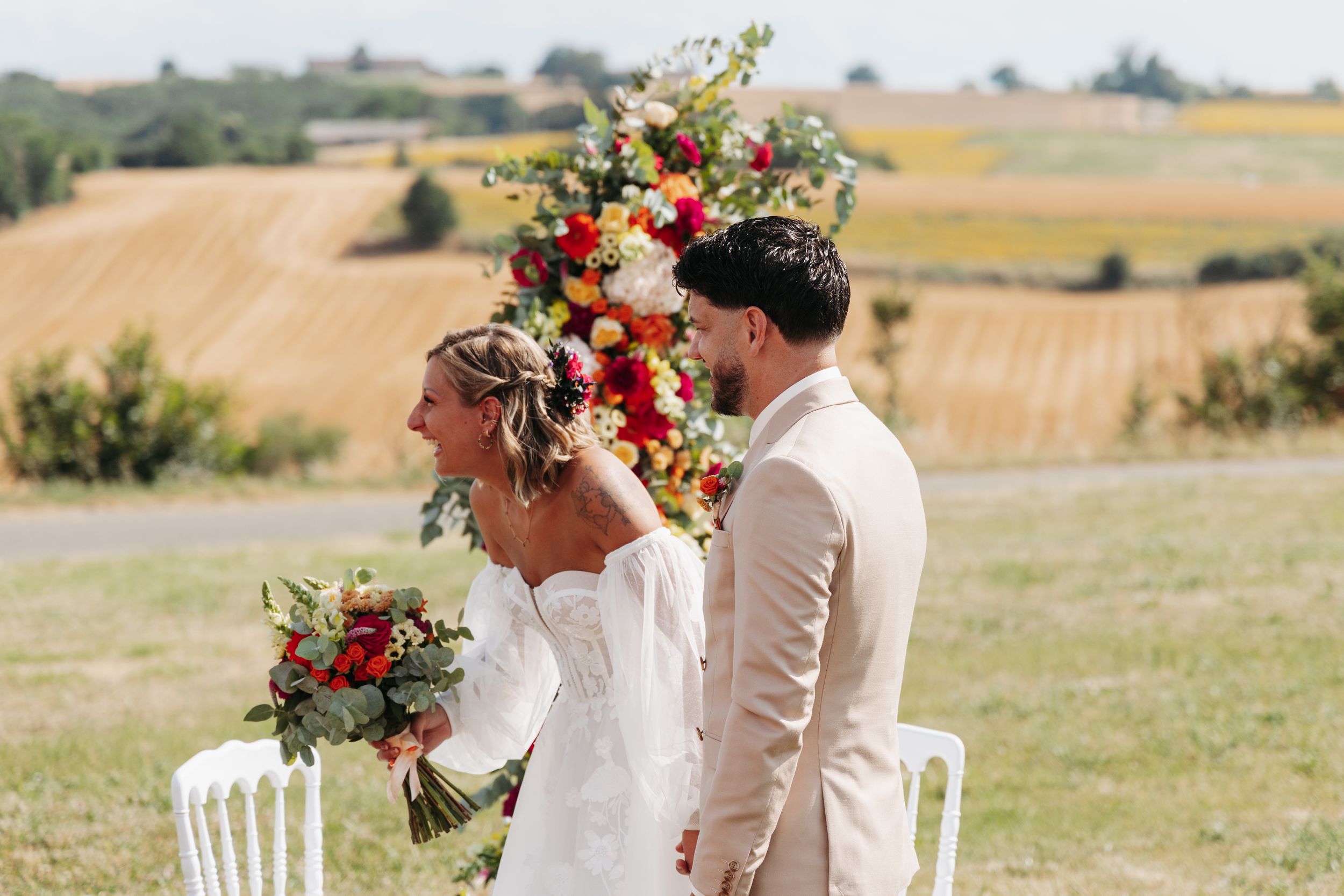 Emotional bride laughs holding bouquet beside groom at colorful floral arch in sunny vineyard.