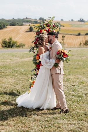 Bride and groom share romantic kiss beside lush floral arch with golden vineyard landscape behind.