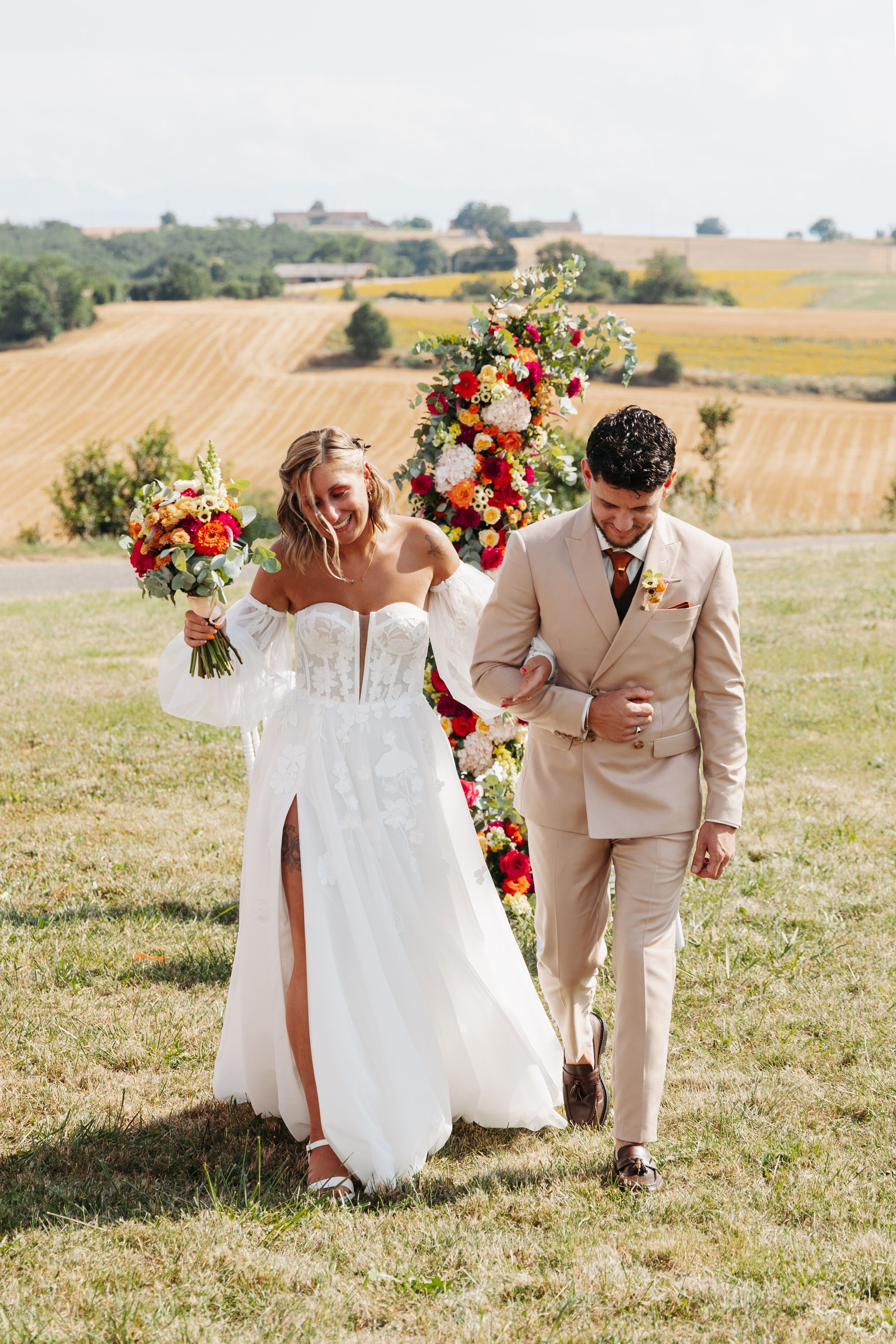 Joyful newlyweds walk arm in arm after ceremony with vibrant bouquet and vineyard backdrop.