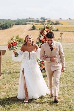 Smiling bride and groom walking after ceremony with colorful floral arch and vineyard backdrop.