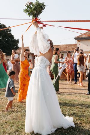 Bride in white gown tossing bouquet with red ribbons to excited wedding guests outdoors.