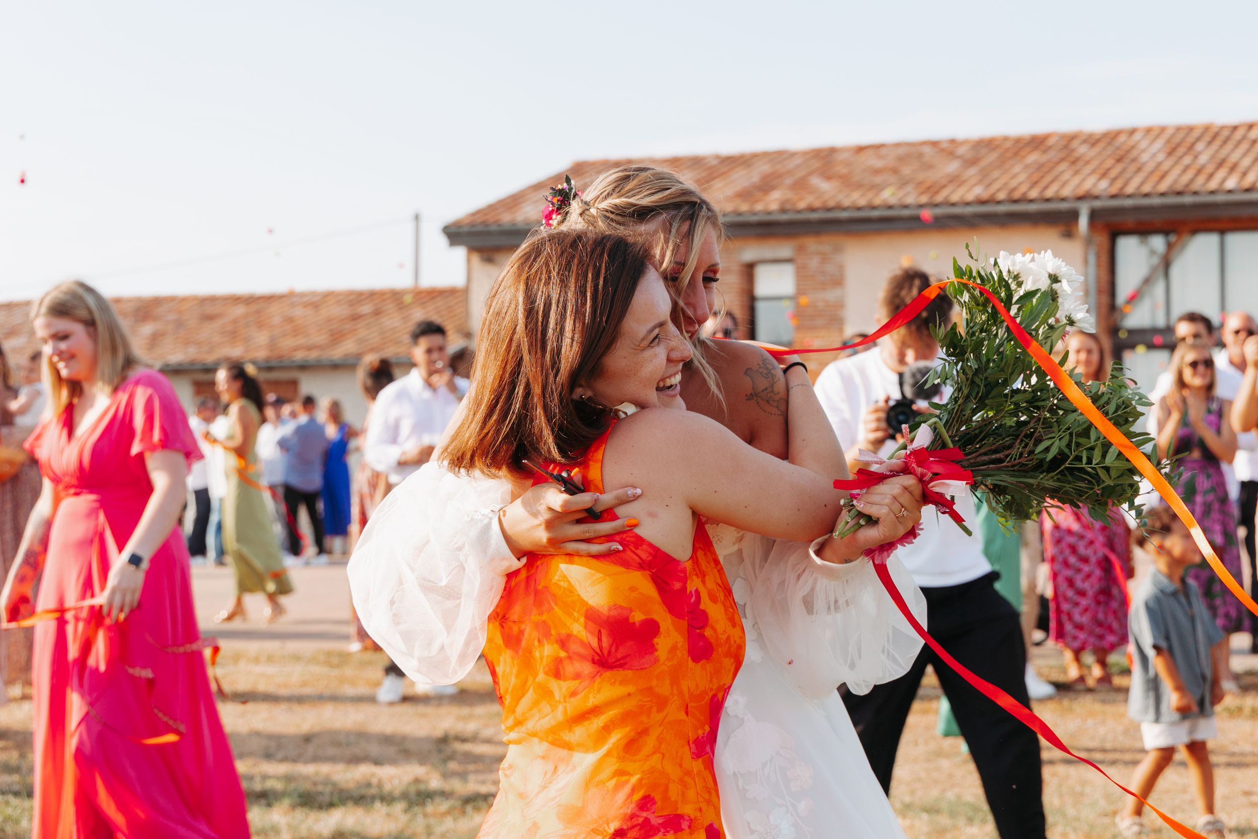 Bride in white gown embracing guest in orange dress during outdoor wedding bouquet toss.