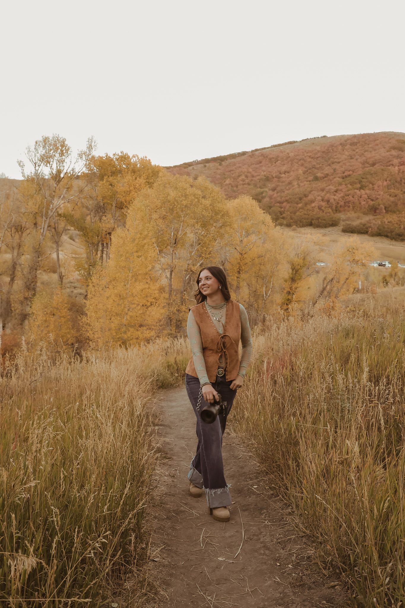 Person walking on dirt trail through tall autumn grasses with golden trees and hills.