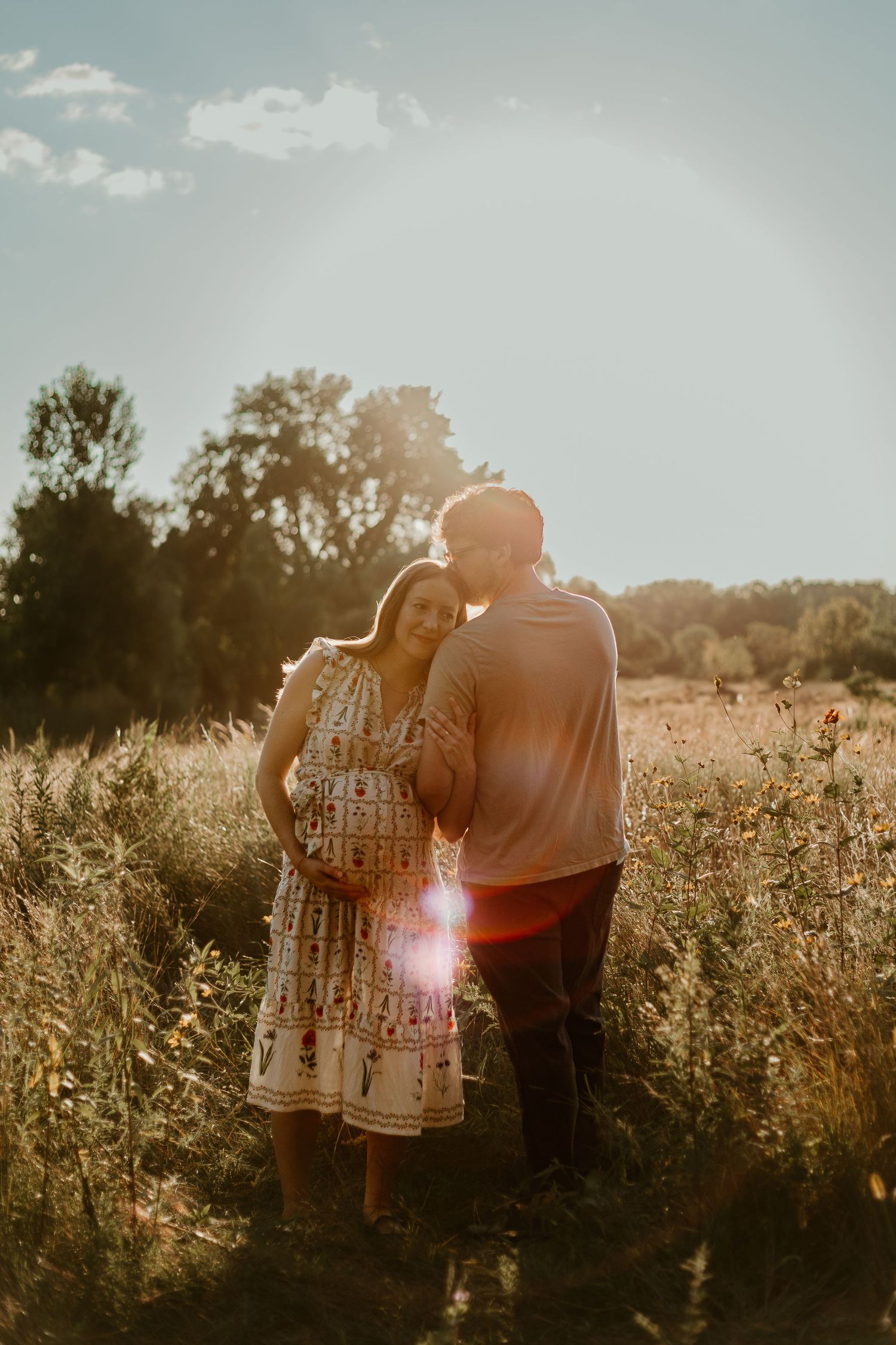 A couple stands together in a sunlit meadow during golden hour as lens flares create dreamy atmosphere.