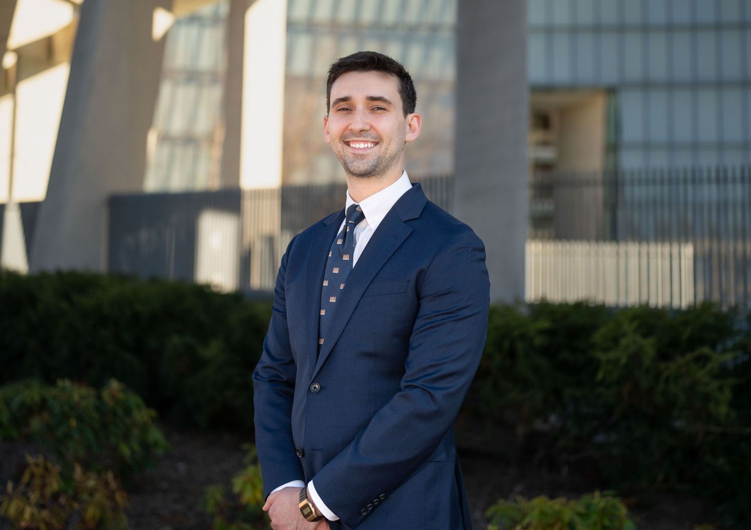 Professional in a navy suit poses with crossed arms in front of a modern office building exterior.