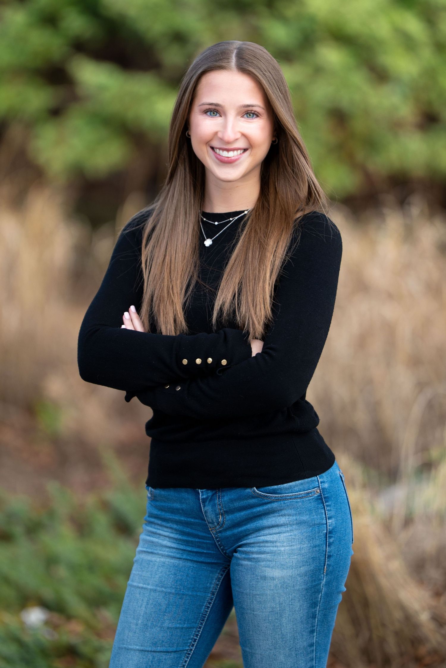 A person in a black sweater and blue jeans poses outdoors with crossed arms and a warm smile.